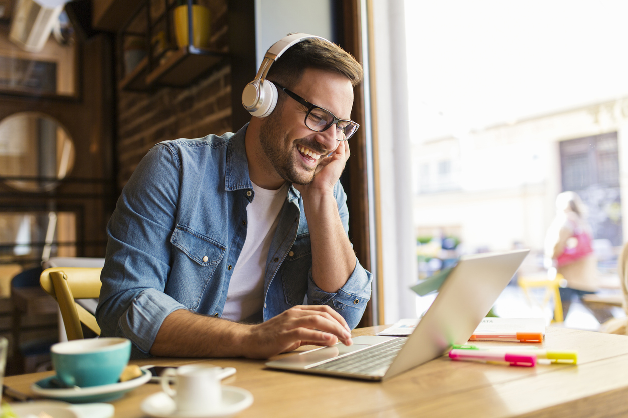 a young man smiling while working at a laptop in a coffee shop