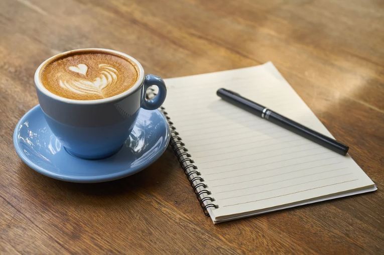 a cup of coffee, a notebook and a pen; placed on a wooden table