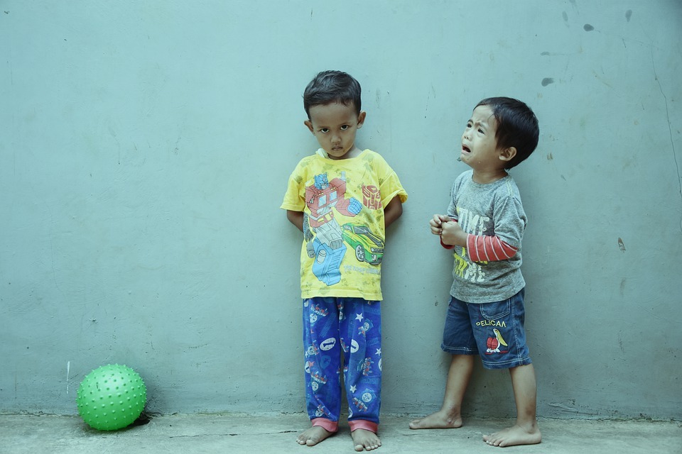 two little boys leaning on a dirty wall, smaller boy crying, taller boy looking angry, green ball on the ground