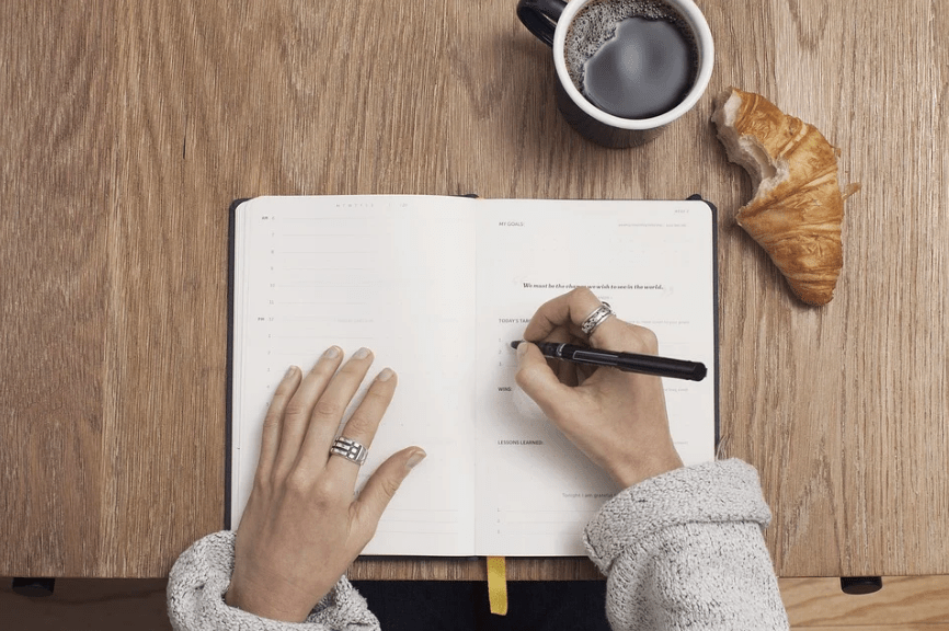 wooden table, cup of black coffee, person writing on the journal, croissant with a bite